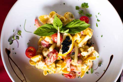 A colorful pasta dish with cherry tomatoes, herbs, and a garnish of basil and caviar, served in a white bowl.