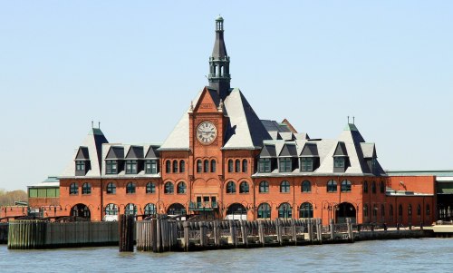 Historic brick building with a clock tower, situated by the water, featuring a peaked roof and surrounding wooden pilings.