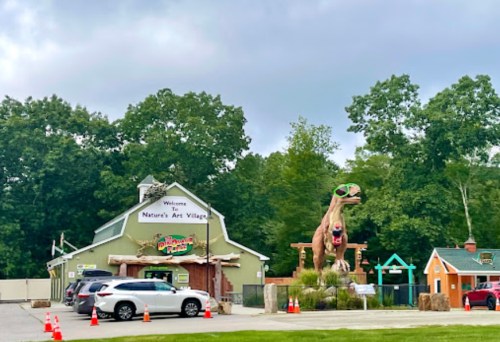 A large dinosaur statue wearing sunglasses stands outside Nature's Art Village, with cars parked nearby and trees in the background.