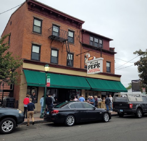 A crowd gathers outside Frank Pepe Pizzeria, a brick building with green awnings and a sign, on a cloudy day.