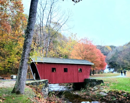 A red covered bridge over a stream, surrounded by autumn trees with colorful foliage. People walk in the background.