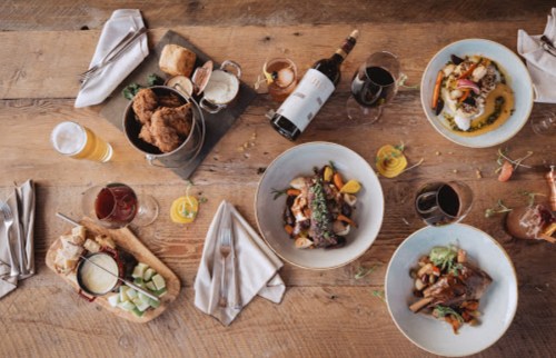 A table spread with various gourmet dishes, drinks, and bread, set on a rustic wooden surface.