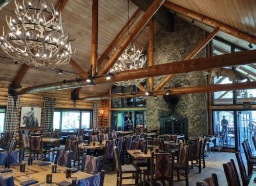 Cozy restaurant interior with wooden beams, antler chandeliers, stone fireplace, and rustic decor.