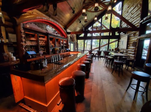 Cozy log cabin bar with a wooden counter, stools, and large windows showcasing a natural view. Warm lighting enhances ambiance.