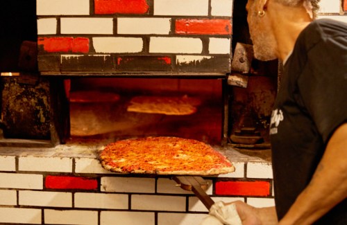 A chef pulls a freshly baked pizza from a brick oven, with red and white brick walls in the background.