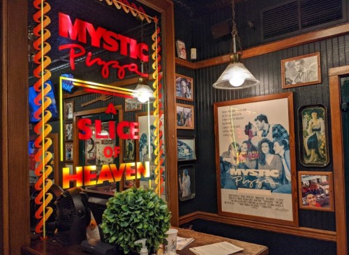 Interior of Mystic Pizza featuring neon signs, movie posters, and a decorative plant on a table.