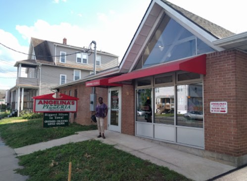 A person stands outside Angelina's Pizzeria, a brick building with a red awning and a sign advertising pizza.