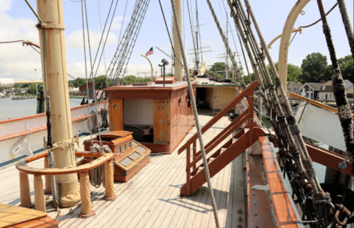 View of a wooden ship's deck with masts, rigging, and a cabin, set against a clear blue sky.