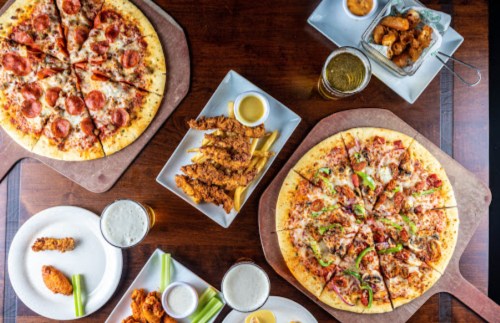 A table spread with pizzas, chicken tenders, celery, dipping sauces, and beers, showcasing a variety of delicious dishes.