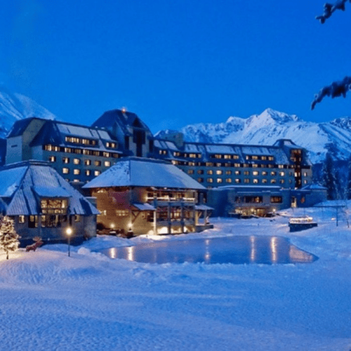 A snowy hotel scene at dusk, surrounded by mountains, with a frozen pond in the foreground and warm lights glowing.
