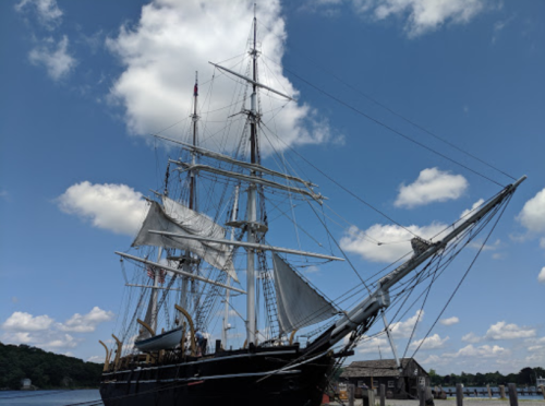 A tall ship with sails raised docked by the water under a blue sky with fluffy clouds.