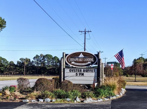 Sign for Chesapeake Landing featuring an oyster buffet at 5 PM, with an American flag and landscaped surroundings.