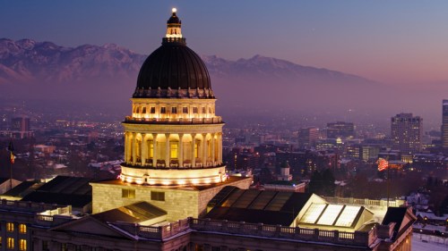 The illuminated dome of a government building at dusk, with mountains and a city skyline in the background.