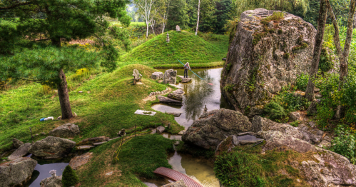 A serene landscape featuring a pond, lush greenery, and a person walking on a bridge among large rocks.