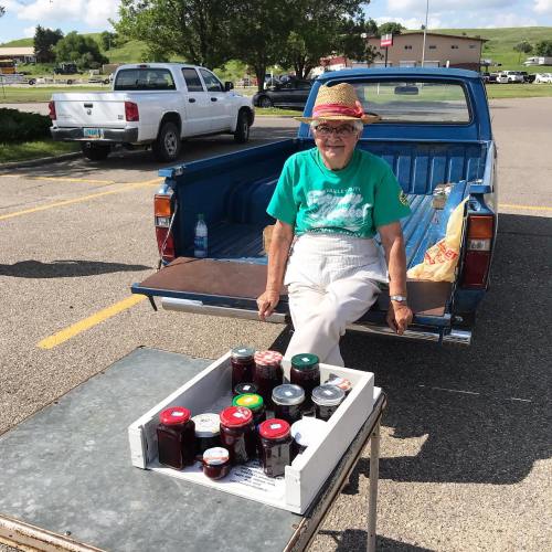 An elderly woman in a straw hat sits by a pickup truck, selling jars of homemade preserves from a table.