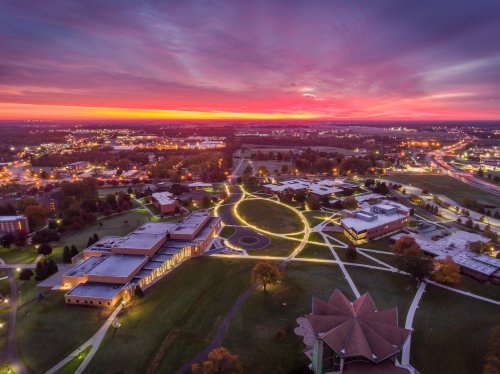 Aerial view of a campus at sunset, with vibrant colors in the sky and illuminated pathways winding through green spaces.