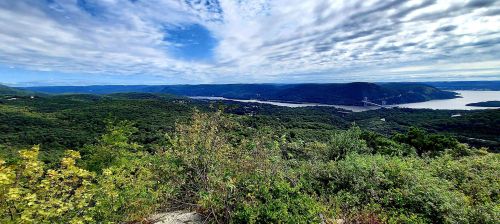 A panoramic view of lush green hills and a river under a cloudy blue sky.