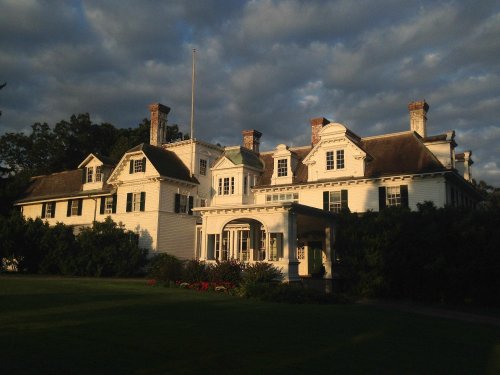 A large, historic white house with multiple chimneys, surrounded by greenery and under a cloudy sky.