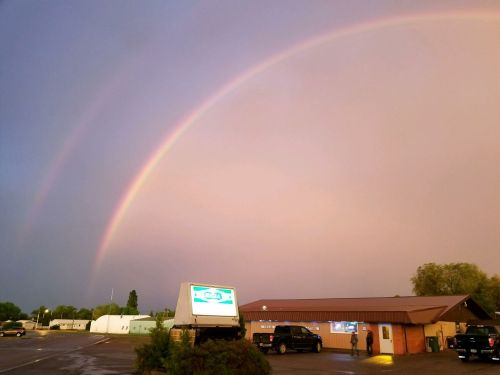 A vibrant double rainbow arcs over a small building and parking lot after a rainstorm.