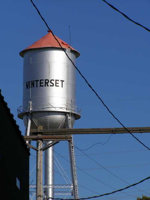 A tall silver water tower with a red roof, labeled "WINTERSET," against a clear blue sky and power lines.