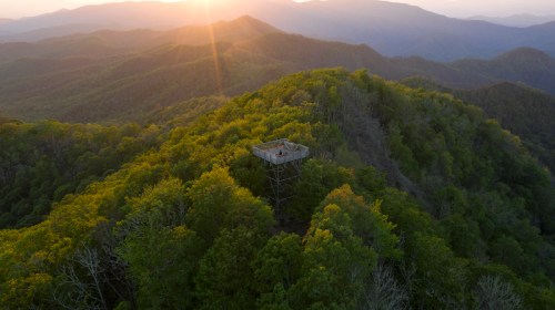 Aerial view of a wooden lookout tower on a lush green mountain, with a sunset illuminating the landscape.