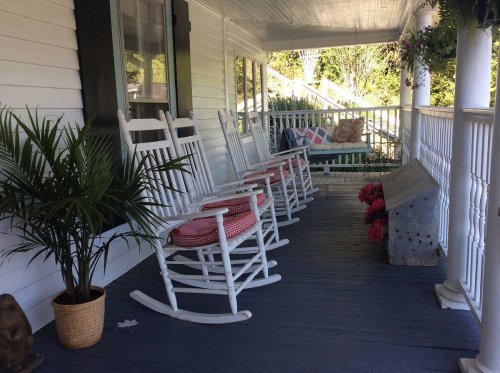 A cozy porch with white rocking chairs, a potted plant, and a swing, surrounded by greenery and a peaceful atmosphere.