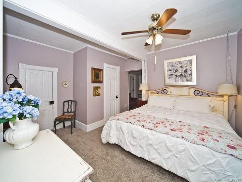 Cozy bedroom with a floral bedspread, ceiling fan, and light purple walls, featuring a chair and decorative vase.