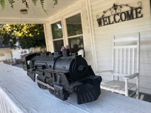A vintage toy train sits on a white railing, with a welcoming porch and greenery in the background.