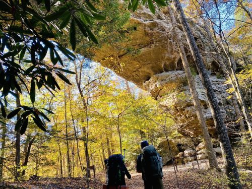 Two hikers with backpacks walk under a large rock formation surrounded by colorful autumn trees.