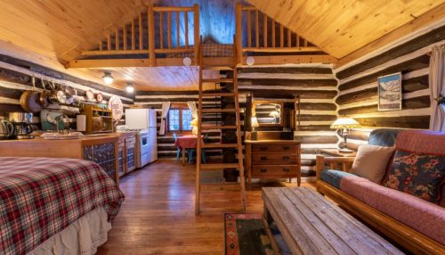 Cozy log cabin interior with a loft, wooden furniture, a kitchen area, and warm lighting.