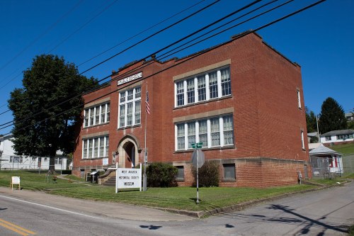 A red brick school building with large windows, an American flag, and a sign in front, set against a clear blue sky.