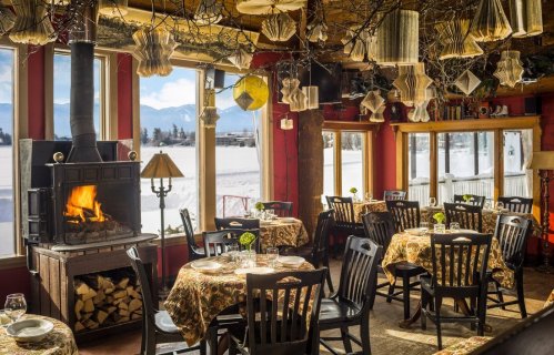 Cozy restaurant interior with tables, a fireplace, and large windows showcasing a snowy landscape outside.