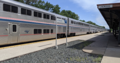 A silver Amtrak train at a station with a sign reading "Lacrosse" and trees in the background.