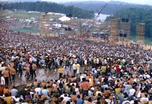 A large crowd of people at a music festival, with a stage in the background and a scenic landscape beyond.