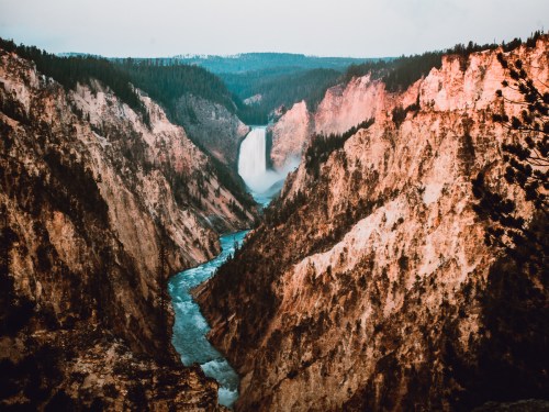 A stunning view of a waterfall cascading into a river, surrounded by rugged cliffs and lush forested mountains.