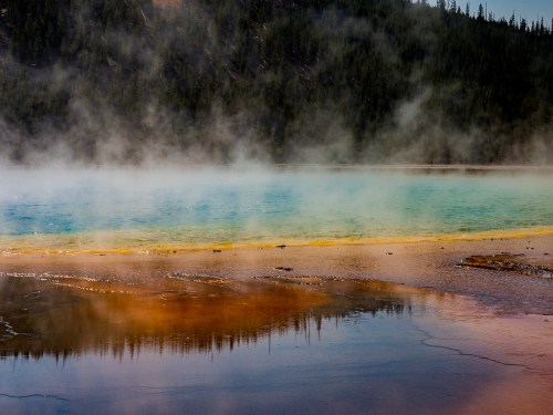 A vibrant hot spring with blue and yellow hues, surrounded by mist and forested hills in the background.
