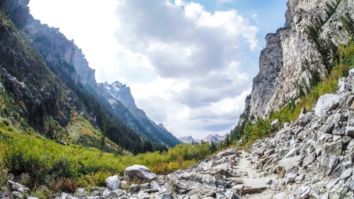 A scenic mountain valley with rocky terrain, lush greenery, and towering cliffs under a partly cloudy sky.