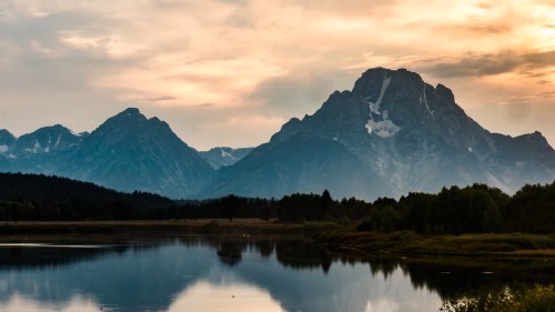 A serene landscape featuring mountains reflected in a calm lake at sunset, with soft clouds in the sky.