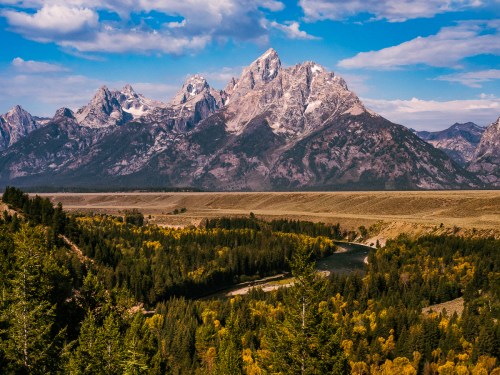 A scenic view of rugged mountains with snow-capped peaks, surrounded by lush forests and a winding river below.