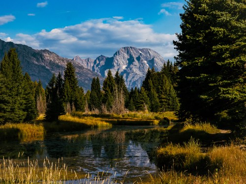 A serene landscape featuring a calm river, lush trees, and majestic mountains under a blue sky with scattered clouds.