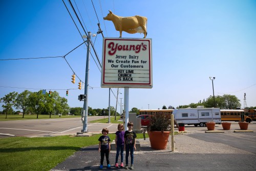 Three children stand in front of Young's Jersey Dairy sign featuring a cow, with a clear blue sky in the background.