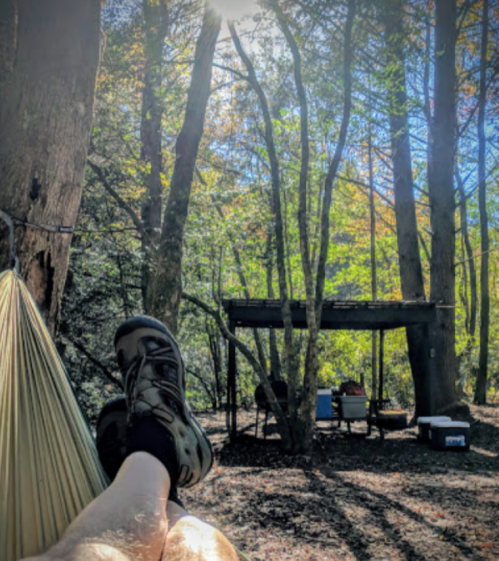 A person relaxes in a hammock surrounded by trees, with a campsite and sunlight filtering through the leaves.