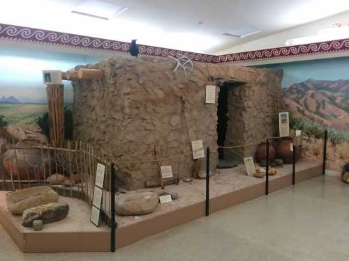 A stone structure resembling a historical dwelling, surrounded by artifacts and informational displays in a museum setting.