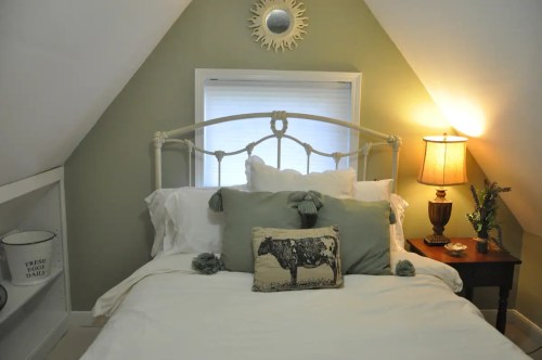 Cozy bedroom with a sloped ceiling, featuring a bed with decorative pillows, a lamp, and a small shelf.