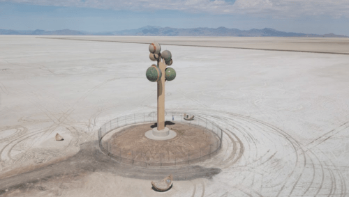 A large, colorful tree sculpture stands on a barren salt flat, surrounded by a circular fence and distant mountains.