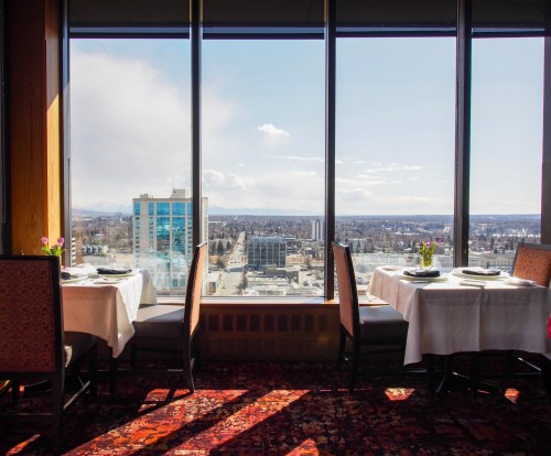 A dining area with tables set for two, overlooking a city skyline through large windows.