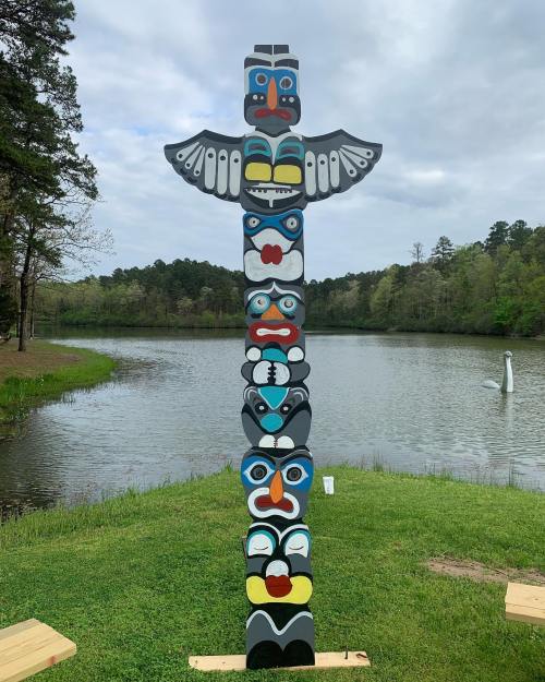A colorful totem pole stands by a serene lake, surrounded by trees and a cloudy sky.