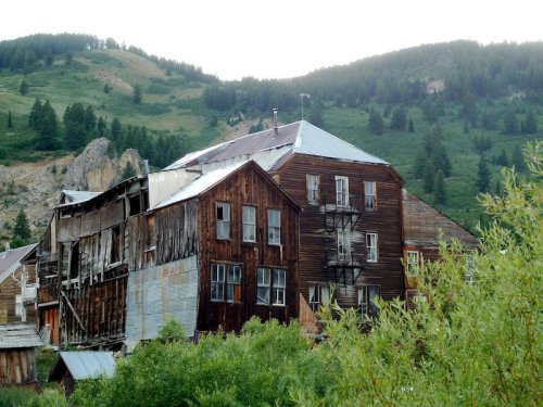 Abandoned wooden building on a hillside, surrounded by greenery and mountains in the background.