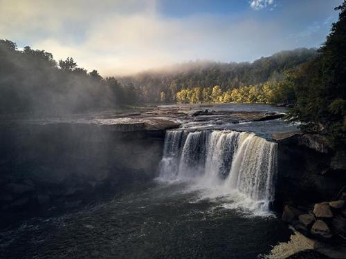 A serene waterfall cascades over rocky cliffs, surrounded by mist and lush green trees under a clear blue sky.