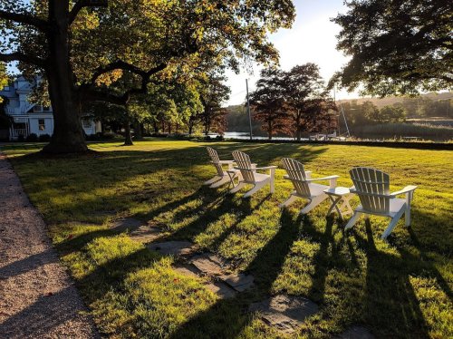 Four white Adirondack chairs on a grassy lawn, casting long shadows in the warm glow of sunset by a calm waterway.